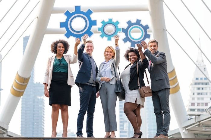 Diverse group of business professionals smiling and holding interconnected gear icons, symbolizing teamwork, collaboration, and organizational efficiency.