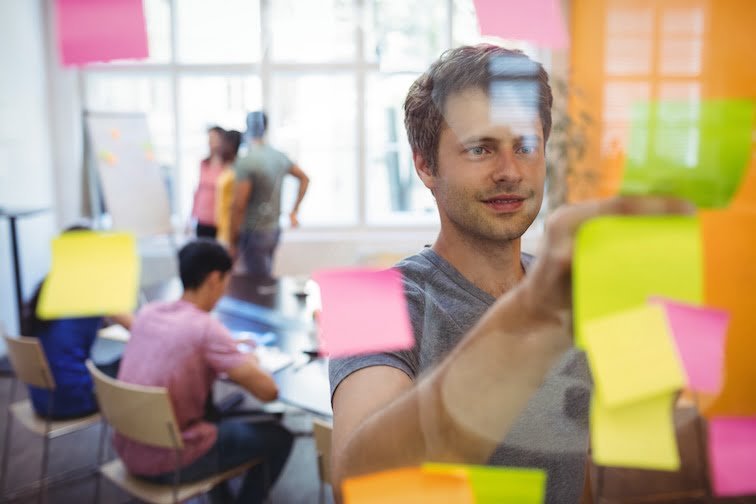 Scrum team member placing colorful sticky notes on a glass wall, with team planning in the background during a sprint session