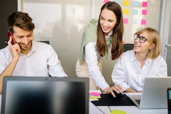 Bir ofiste bir erkek telefonla konuşuyor ve bir kadın diğer kadına bilgisayarda bir şey gösteriyor. İnsanlar gülümsüyorlar.