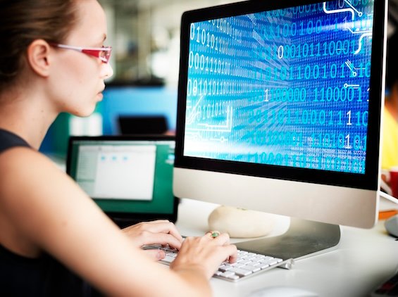 Woman working at a desktop computer displaying binary code, representing software development, programming, or data analysis in a tech-focused environment.