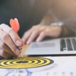 Bir iş insanı, hedef tahtasının tam merkezine kırmızı dart oku yerleştiriyor. Arka planda bir dizüstü bilgisayar ve iş grafiklerinin yer aldığı belgeler bulunuyor. Görsel, stratejik hedef belirleme, odaklanma ve iş başarısına ulaşma kavramlarını simgeliyor.