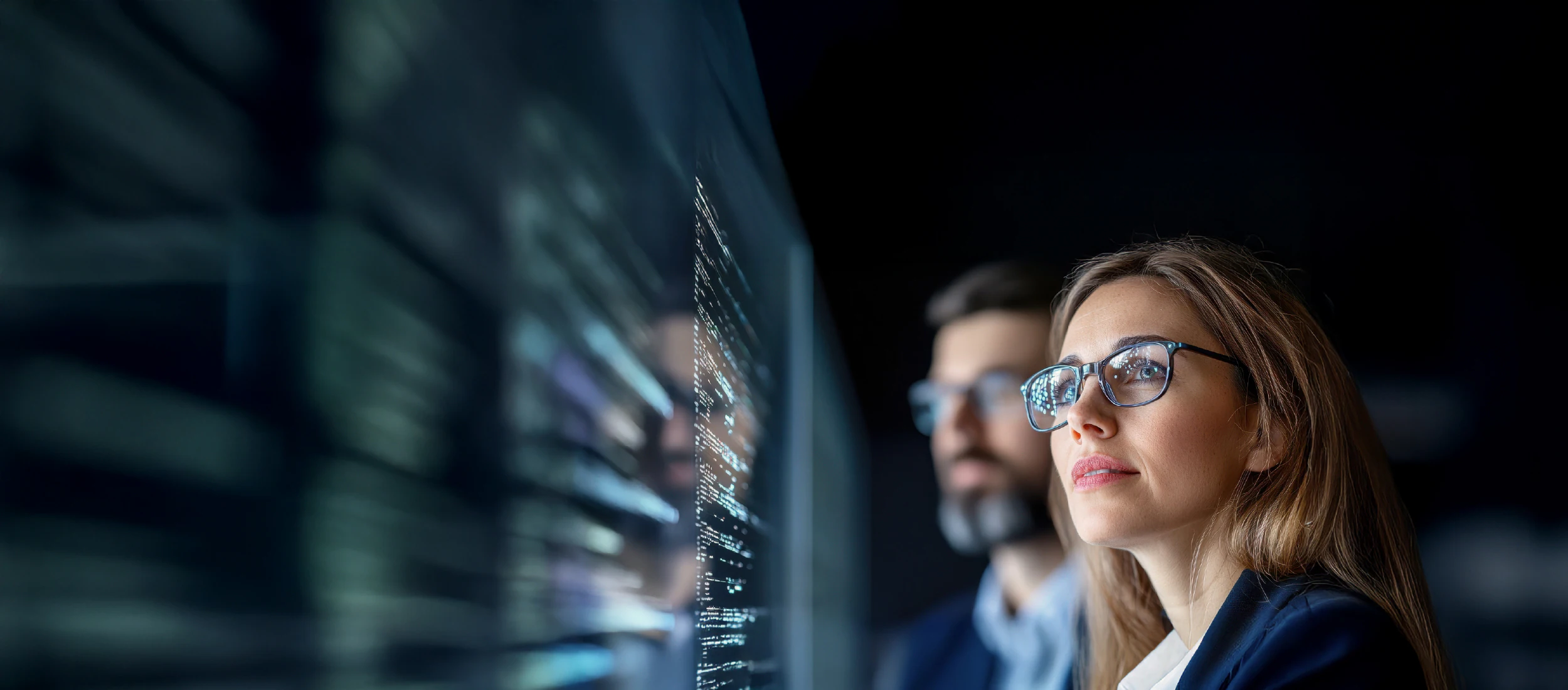 Gözlüklü bir kadın ve arkasında bulanık şekilde görünen bir erkek, karanlık bir ortamda dijital veri ekranına odaklanmış halde yapay zekâ ve yazılım verilerini inceliyor. Ekranda yansıyan ışıklar, kod satırlarını temsil ediyor. Kurumsal teknoloji dönüşümü, veri analitiği ve yapay zekâ kullanımını simgeleyen modern bir sahne.
